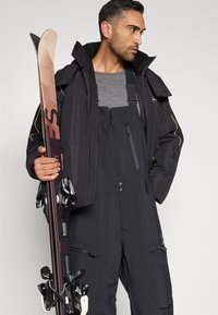 Μαύρο χειμερινό μπουφάν σκι με ψηλό γιακά, συνδυασμένο με μαύρα παντελόνια εργασίας τύπου σαλοπέτα. Το μοντέλο κρατά ένα ζευγάρι σκι με δεσίματα, παρουσιάζοντας μία λεία υφή.