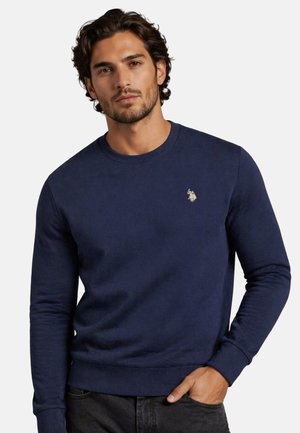Man wearing a navy blue long-sleeve sweatshirt with a small logo on the chest, standing with one hand in his pocket.