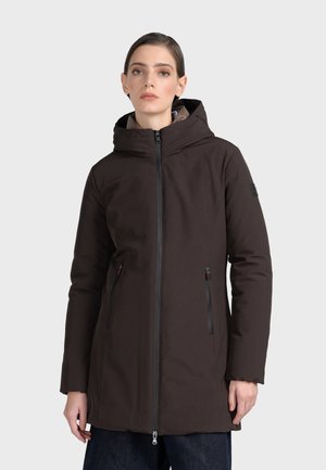 Femme portant un manteau d'hiver à capuche marron foncé avec fermeture éclair devant et poches, debout devant un fond clair uni.