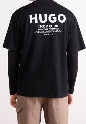 Personne portant un t-shirt noir de marque HUGO avec un texte blanc et un pantalon beige, vue de dos.