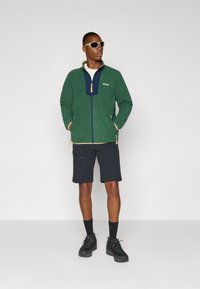 Veste en polaire verte avec une fermeture éclair navy à l'avant, accents en beige, short navy, chaussures de randonnée noires et lunettes de soleil claires. Design simpliste.