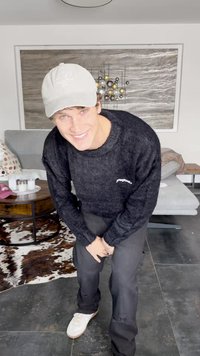Black knitted sweater with a round neckline, paired with black distressed pants and white sneakers. A light gray cap completes the look.