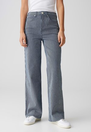 Personne portant un jean taille haute à jambes larges bleu et blanc à fines rayures, avec des baskets blanches et un top blanc sans manches.
