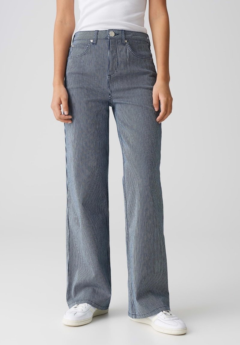 Personne portant un jean taille haute à jambes larges bleu et blanc à fines rayures, avec des baskets blanches et un top blanc sans manches.