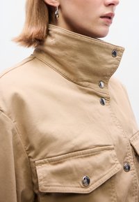 Μπεζ μπουφάν με ψηλό γιακά, κουμπώματα με τρουκ και δύο μπροστινές τσέπες. Λείο ύφασμα με διακριτική υφή και ορατές ραφές.