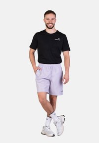 T-shirt nera di cotone con logo, pantaloni corti viola chiaro, vita con cordino, e scarpe da ginnastica argentate con calzini bianchi a righe nere.