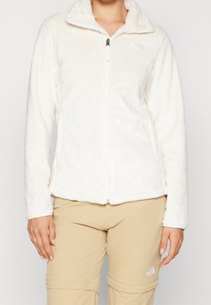 Personne portant une veste polaire blanche avec zip frontal et un pantalon convertible beige, debout devant un fond uni.
