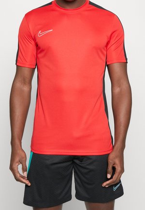 Man draagt een rood Nike sportshirt met korte mouwen en zwarte strepen aan de zijkanten en zwarte sportshorts met turquoise accenten.