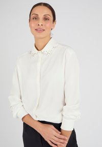Blusa blanca con un cuello decorativo adornado con tachuelas, mangas largas y cierre de botones en la parte delantera. Tela suave y corte relajado, adecuada para combinar en capas.