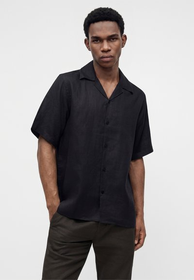 Jeune homme portant une chemise noire à manches courtes boutonnée et un pantalon foncé, debout avec une main dans la poche sur un fond blanc uni.