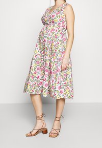 Vestido de maternidad sin mangas y con estampado floral, con escote en V, cintura ceñida y falda acampanada. Los colores incluyen rosa, amarillo y verde. Sandalias de tiras marrones.