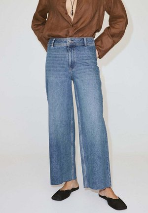 Person trägt hoch taillierte, weit geschnittene blaue Jeans, braunes langärmeliges Shirt, schwarze flache Slipper und eine schwarze Anhängerkette.
