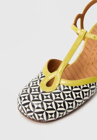 Sort-hvid sko med geometrisk mønster, gul rem og gul fremhævelse. Rundt tådesign med udskæring foran.