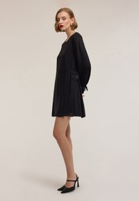 Abito nero con maniche lunghe, caratterizzato da dettagli con perline sui polsini. Il tessuto è liscio, dalla silhouette morbida e lunghezza corta.