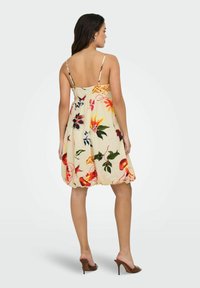 Femme portant une robe sans manches, jusqu'aux genoux, de couleur crème avec des motifs floraux colorés et des chaussures à talons hauts ouvertes marron, se tenant devant un fond uni.