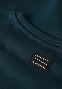 Tessuto verde scuro con una piccola patch rettangolare nera vicino al colletto che recita "CREA IL TUO DOMANI" in lettere maiuscole bianche.