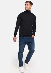 Schwarzer Rollkragenpullover mit langen Ärmeln, kombiniert mit blauen Jeans und weißen Sneakers. Sanfte Textur, taillierte Passform.