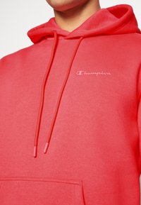 Sudadera roja hecha de tela suave, con un bolsillo frontal, cordones y un pequeño logotipo blanco en el pecho. Textura suave.