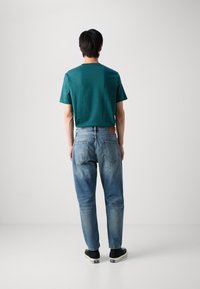 Teal cotton t-shirt, relaxed fit, paired with faded blue denim jeans featuring back pocket stitching and brown leather patch, worn with black sneakers.