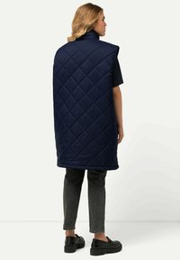 Gilet trapuntato blu navy con colletto alto, design senza maniche e motivo a cucitura diagonale, abbinato a pantaloni grigio scuro e scarpe nere.