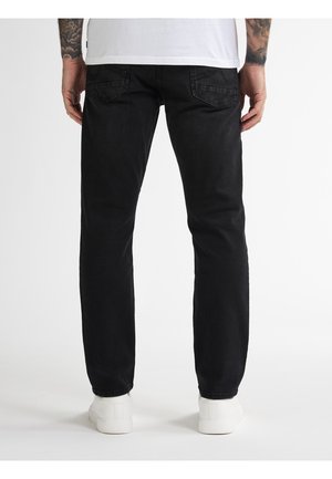 Mustat farkut suoralahkeisella leikkauksella, joissa on kaksi takataskua ja minimaalinen ompelu. Päällä valkoiset lenkkarit.