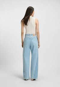 Kobieta stoi tyłem, ubrana w bezrękawnik z koronką w kolorze kremowym oraz jasnoniebieskie spodnie z wysokim stanem i szerokimi nogawkami, do tego beżowe obcasy.