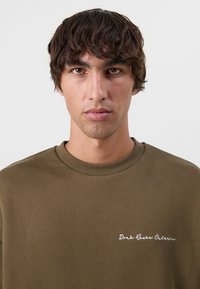 Olivengrønn sweatshirt laget av bomull, med rund hals, ribbede mansjetter og brodert tekst på venstre side.