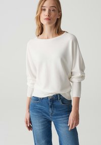 Weißer, weicher Pullover mit lockerer Passform und weitem Ausschnitt, kombiniert mit mittelblauen geraden Jeans. Der Pullover hat lange Ärmel und gerippte Bündchen.