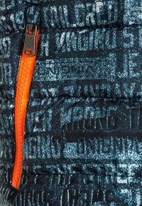 Svart puffjacka med ett abstrakt blått mönster, som har en orange dragkedja och textil med struktur. Inkluderar synliga sömmar.