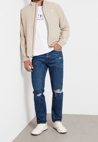 Beżowa kurtka na zamek z wysokim kołnierzem, nałożona na białą koszulkę z nadrukiem, zestawiona z podniszczonymi niebieskimi dżinsami i białymi tenisówkami.