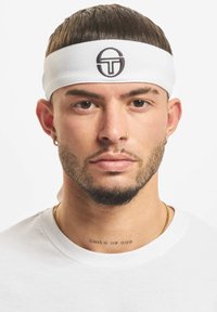 Bílá sportovní čelenka s texturovaným povrchem a černým kulatým logem uprostřed. Model nosí jednobarevné bílé tričko.