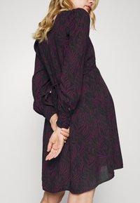 Vestido preto com um design fluido, apresentando um padrão de onda roxa, mangas longas e bufantes, e um detalhe franzido no pulso.
