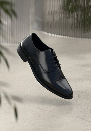 Zwarte gepolijste leren nette schoen met veters, zwevend weergegeven boven een lichte betonnen vloer met een wazige achtergrond.