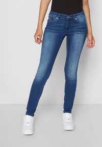 Jeans ajustados en denim azul. Presentan una cintura de altura media, cinco bolsillos, un sutil desgaste y una textura suave. Combinados con zapatillas blancas.