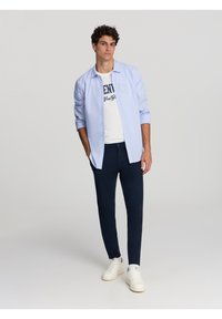 Camicia azzurra chiara a maniche lunghe indossata sopra una t-shirt bianca con grafica, abbinata a pantaloni blu scuro e sneaker bianche. Elementi di design semplici e moderni.