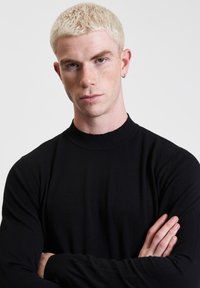 Maglione nero a collo alto con maniche lunghe, texture morbida, design aderente, indossato da una persona con capelli biondi corti e orecchio forato.