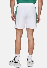 Pantaloni corti bianchi con pannelli laterali verdi e blu, cintura elastica e tessuto testurizzato. Indossati con sneaker nere e bianche e calze bianche.