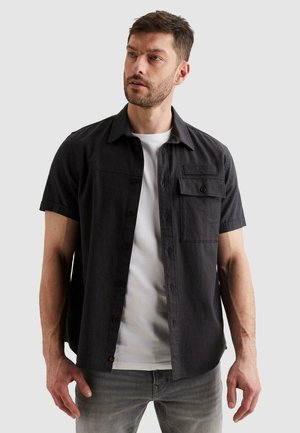 Man draagt een zwart overhemd met korte mouwen over een wit T-shirt en grijze jeans, kijkt iets naar links met een neutrale uitdrukking.