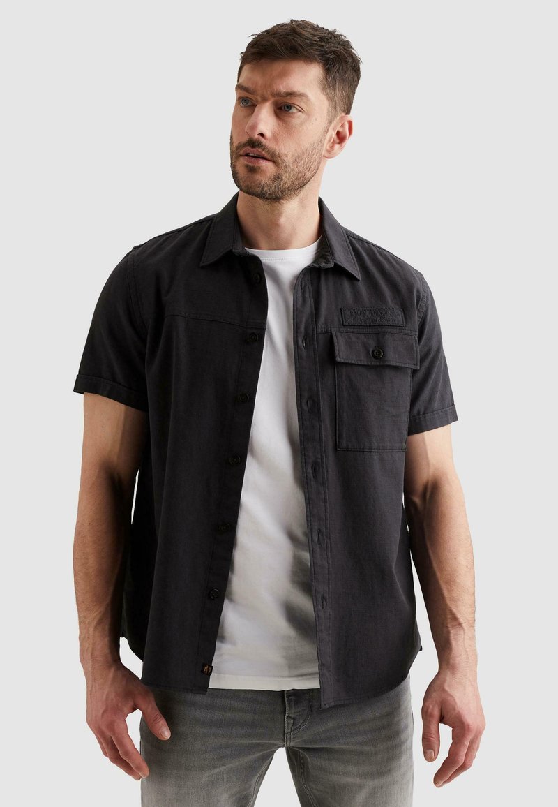 Homme portant une chemise noire à manches courtes boutonnée sur un t-shirt blanc et un jean gris, regardant légèrement vers la gauche avec une expression neutre.