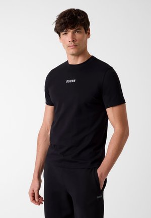 Homme debout avec une main dans la poche, portant un t-shirt noir Guess et un pantalon noir assorti, devant un fond clair uni.