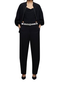 Zwarte bomberjack over een zwart topje, high-waisted zwarte broek met een zilveren glitterriem, en zwarte puntige flats. Glad materiaal en ontspannen pasvorm.