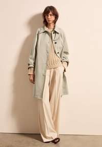 Lichtgroene trenchcoat met een kraag en zwarte knopen, gedragen over een beige gebreide sweater en beige wijde broek. Glad van textuur.