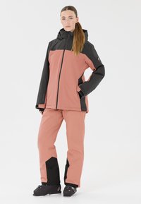 Rosa und schwarze Skijacke mit Frontreißverschluss und Kapuze, kombiniert mit passenden rosa Skihosen mit schwarzen Akzenten und Taschen. Schwarze Skischuhe.