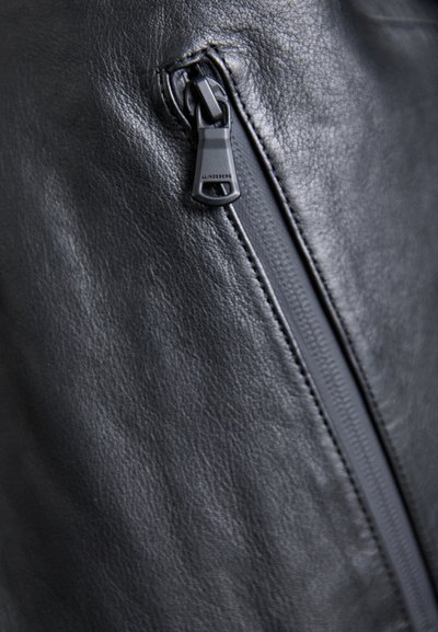 Tecido de couro preto com um detalhe em zipper no lado direito, com uma aba para puxar. A textura é lisa e ligeiramente brilhante.