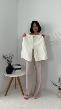 Nainen, jolla on mustat hiukset, pitää valkoisia denim-shortsseja minimalistisessa huoneessa, jossa on beiget housut, pyöreä pöytä ja koristeellisia oksia maljakoissa.