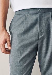 Graue, maßgeschneiderte Hose aus strukturiertem Stoff, mit einer einzelnen vorderen Falte und einem Knopfverschluss am Bund. Hand in der Nähe der Tasche positioniert.