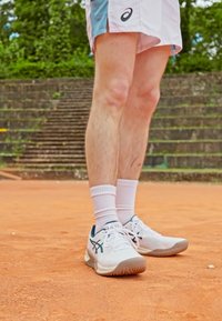 Pantofi sport albi cu un logo albastru, talpă texturată și accente roz. Purtați cu șosete albe, stând pe un teren de tenis din argilă.