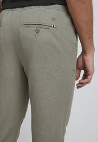 Jasnozielone spodnie o oliwkowym odcieniu, wykonane z fakturowanej tkaniny; posiadają dwie tylne kieszenie z guzikami i subtelne logo u góry. Widoczna elastyczna talia.