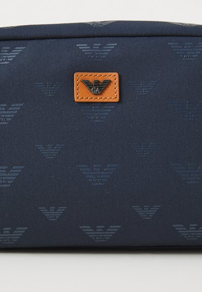 Marineblå stoffpose med utstanset ørn logo mønster. Har en brun lærtag med utstanset merkeinitialer. Glatt tekstur.
