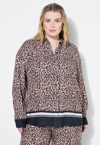 Frau trägt ein langärmliges, geknöpftes Hemd mit Leopardenmuster und einem schwarz-weiß gestreiften Saum, passende Hosen, steht vor einem weißen Hintergrund.